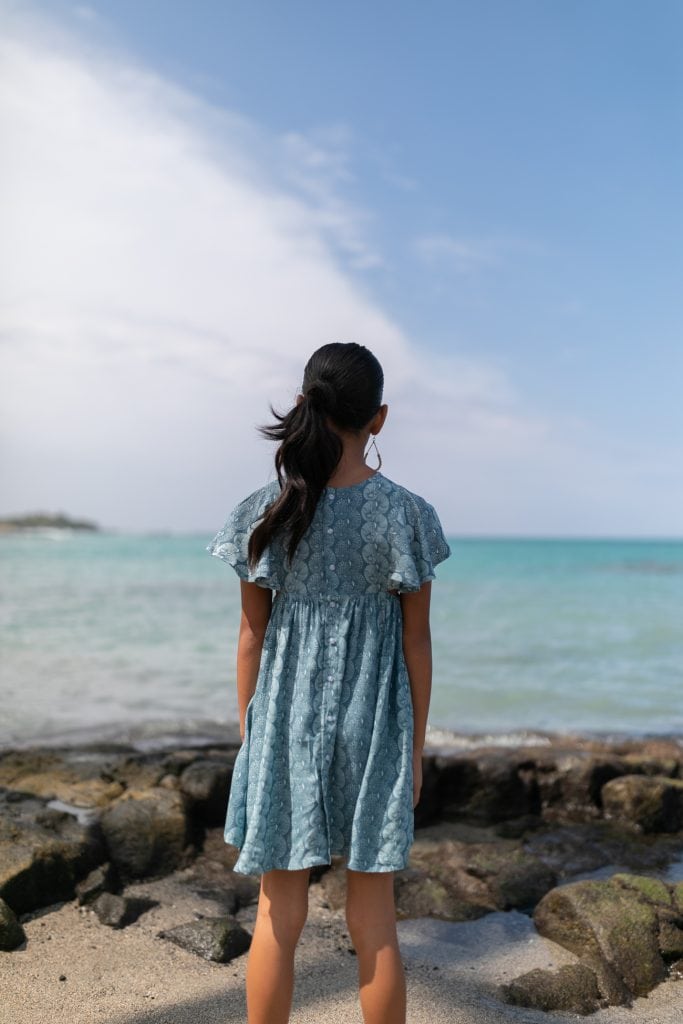 Girl model wearing a Keiki Kailani Dress in a Lei Kupee Print and Real Teal/Blue Tint Color - Back View